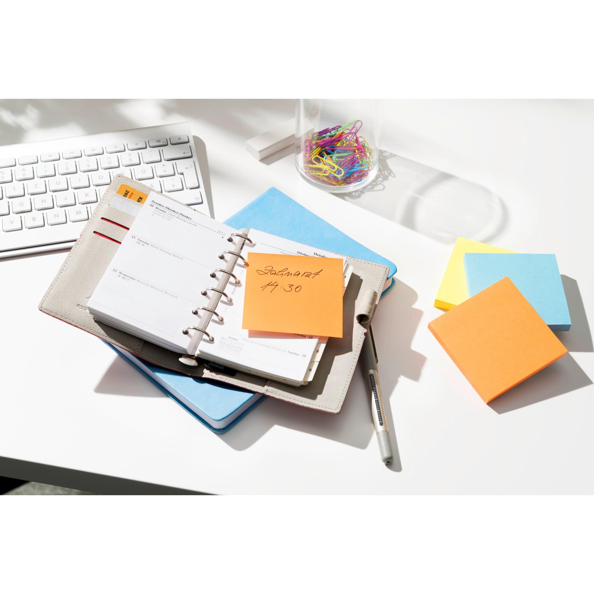 Ein Planer mit einem Stift liegt auf einem Schreibtisch neben einer Tastatur, Büroklammern und einem Stapel Post-it® Notes (76 mm x 76 mm, 100% PEFC) der 3M Deutschland GmbH. Ein orangefarbener Zettel mit der Aufschrift "Zahnarzt 4:30" ist an dem Planer befestigt.