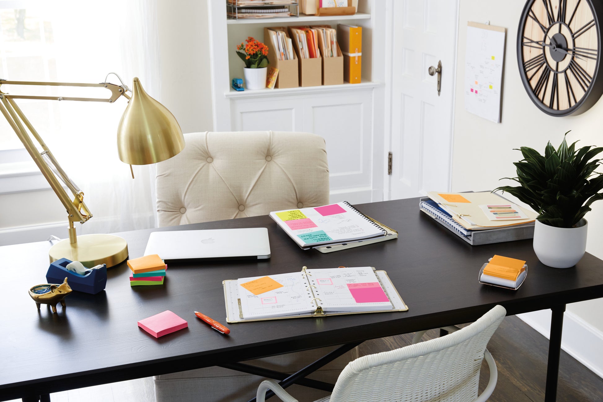 Ein aufgeräumtes Heimbüro mit schwarzem Schreibtisch zeigt einen Laptop, Planer, ein 3M Deutschland GmbH Post-it® Super Sticky Notes 654-6SS-PPNK Paket (rosa, 76x76mm), Lampe, Stifthalter, Pflanze und Dekoration. Im Hintergrund befinden sich Regale, Akten und eine große Wanduhr.