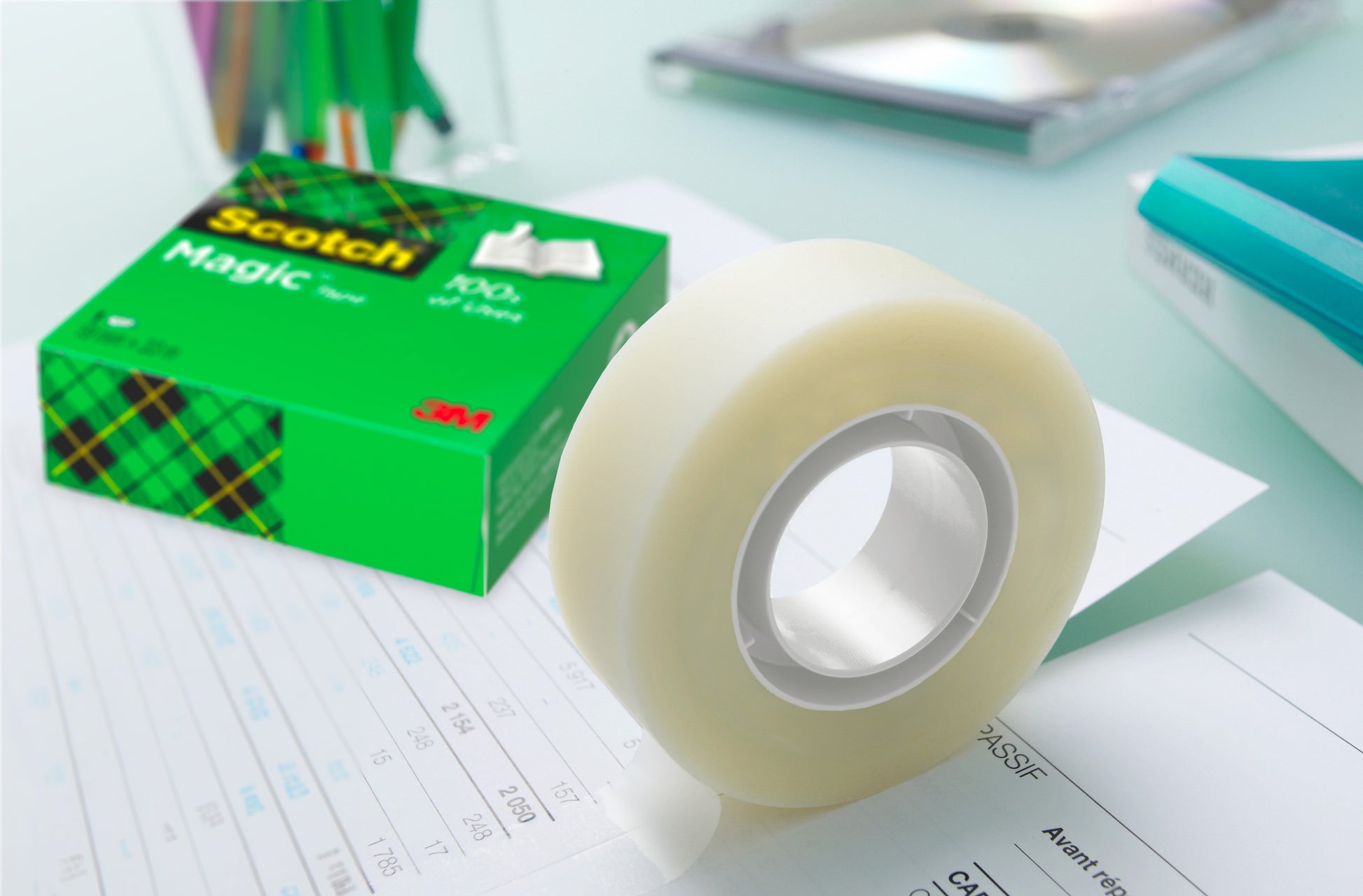 Eine Packung Scotch® Magic™ Unsichtbares Klebeband (8 Rollen, 19 mm x 33 m) von 3M Deutschland GmbH liegt auf einem Dokument, die grüne Schachtel daneben. Stifte, ein Notizbuch und andere Büromaterialien erscheinen unscharf im Hintergrund.