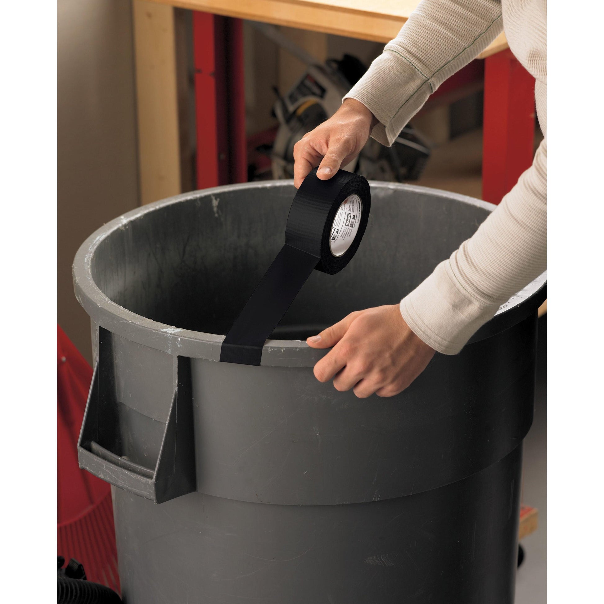 Eine Person klebt einen Streifen Scotch® Extremium™ Universal Klebeband (3 m x 19 mm) von 3M Deutschland GmbH über die Oberseite eines großen grauen Mülleimers und befestigt ihn an den gegenüberliegenden Seiten des Randes. Im Hintergrund sind eine Werkbank und Werkzeuge zu sehen.