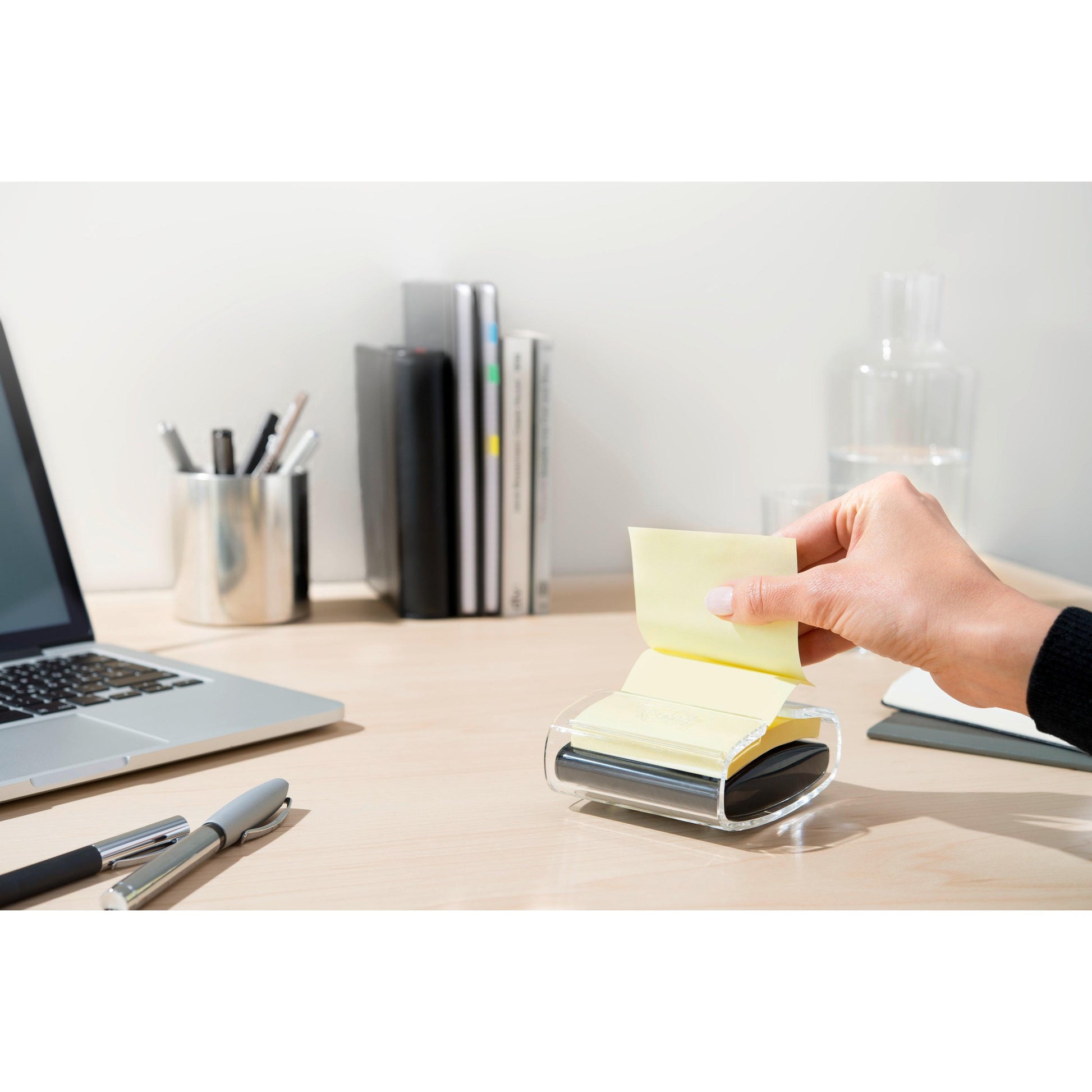 Eine Hand zieht eine gelbe Post-it® Super Sticky Z-Note (76x76 mm) aus dem schwarzen PRO Post-it® Z-Notes Dispenser der 3M Deutschland GmbH auf einem aufgeräumten Schreibtisch mit Laptop, Stiften, Büchern, Glaskaraffe und Tasse, die ordentlich im Hintergrund stehen.