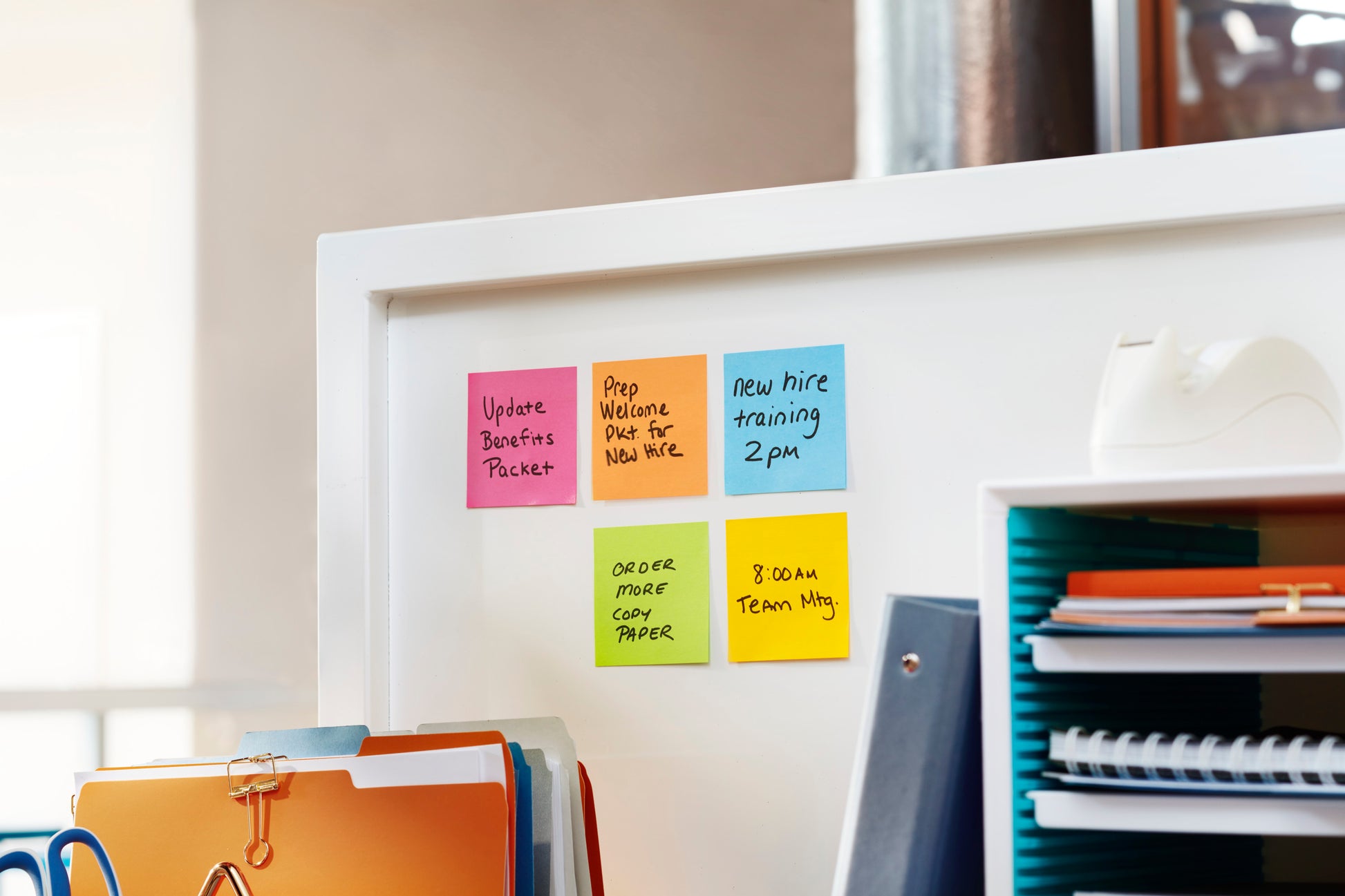 Auf dem Schreibtisch stehen die Post-it® Super Sticky Notes (76 mm x 76 mm, 90 Blatt/Block) der 3M Deutschland GmbH in sechs Farben auf einem weißen Bürotrenner, daneben ein durchsichtiger Klebebandabroller und geordnete Ordner und Notizbücher.