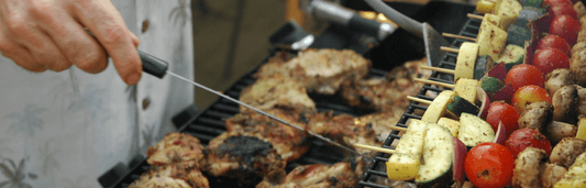 Person grillt Fleischstücke und Gemüsespieße auf einem Grill, hält dabei eine Grillzange über dem Rost in Nahaufnahme