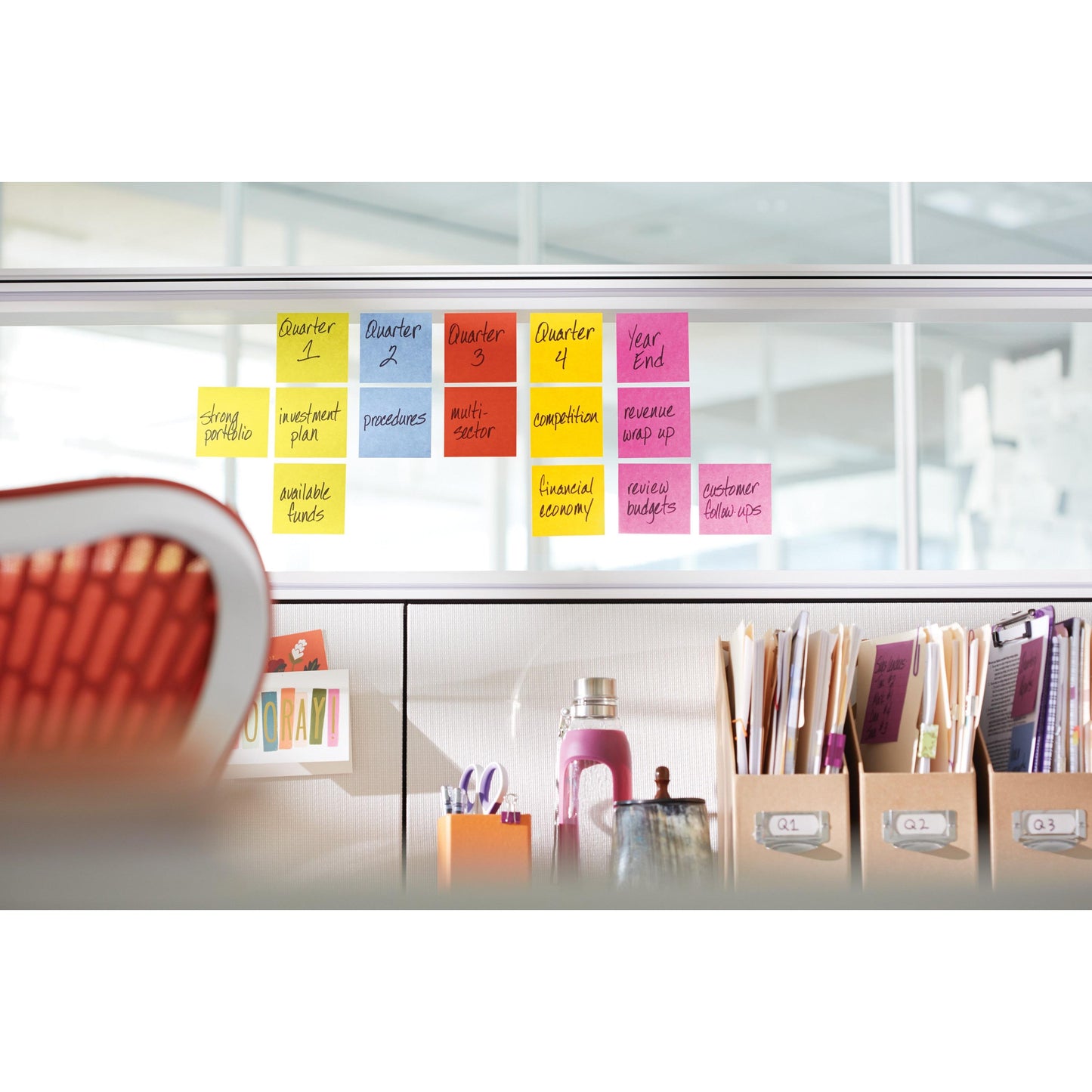An office scene with Post-it® Super Sticky Notes (76 mm x 76 mm, 90 sheets/block, PEFC) from 3M Deutschland GmbH, colorfully arranged on a glass wall for quarterly tasks. In front: folders, documents, a pink bottle, and a red chair.