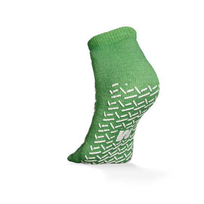 Die ECO-PLUS Anti-Rutsch Einwegsocken beidseitig beschichtet von AMPri Handelsgesellschaft mbH ist eine grüne Knöchelsocke mit weißen rutschfesten Griffen, dargestellt auf einem erhöhten Fuß vor weißem Hintergrund - ideal für medizinische Bereiche.
