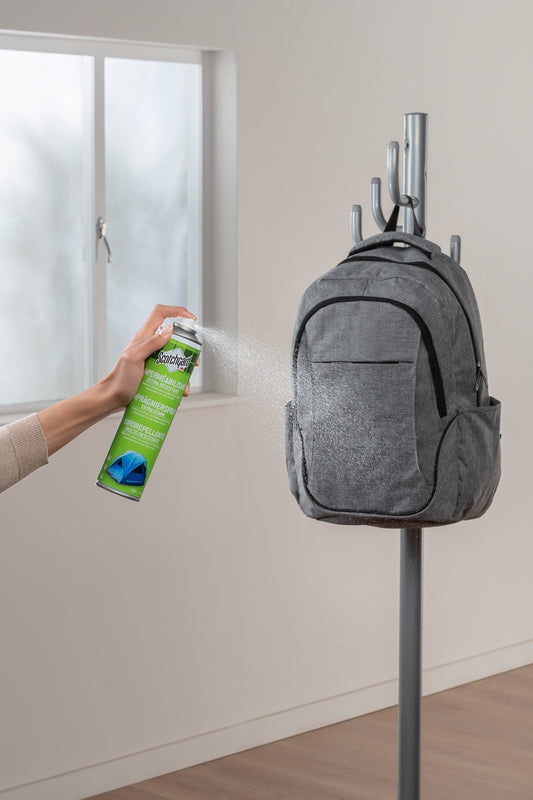 Eine Person sprüht Scotchgard™ Imprägnierspray extra stark (400ml, 1 Dose) von 3M Deutschland GmbH auf einen grauen Rucksack, der an einem Kleiderständer in der Nähe eines Fensters hängt.
