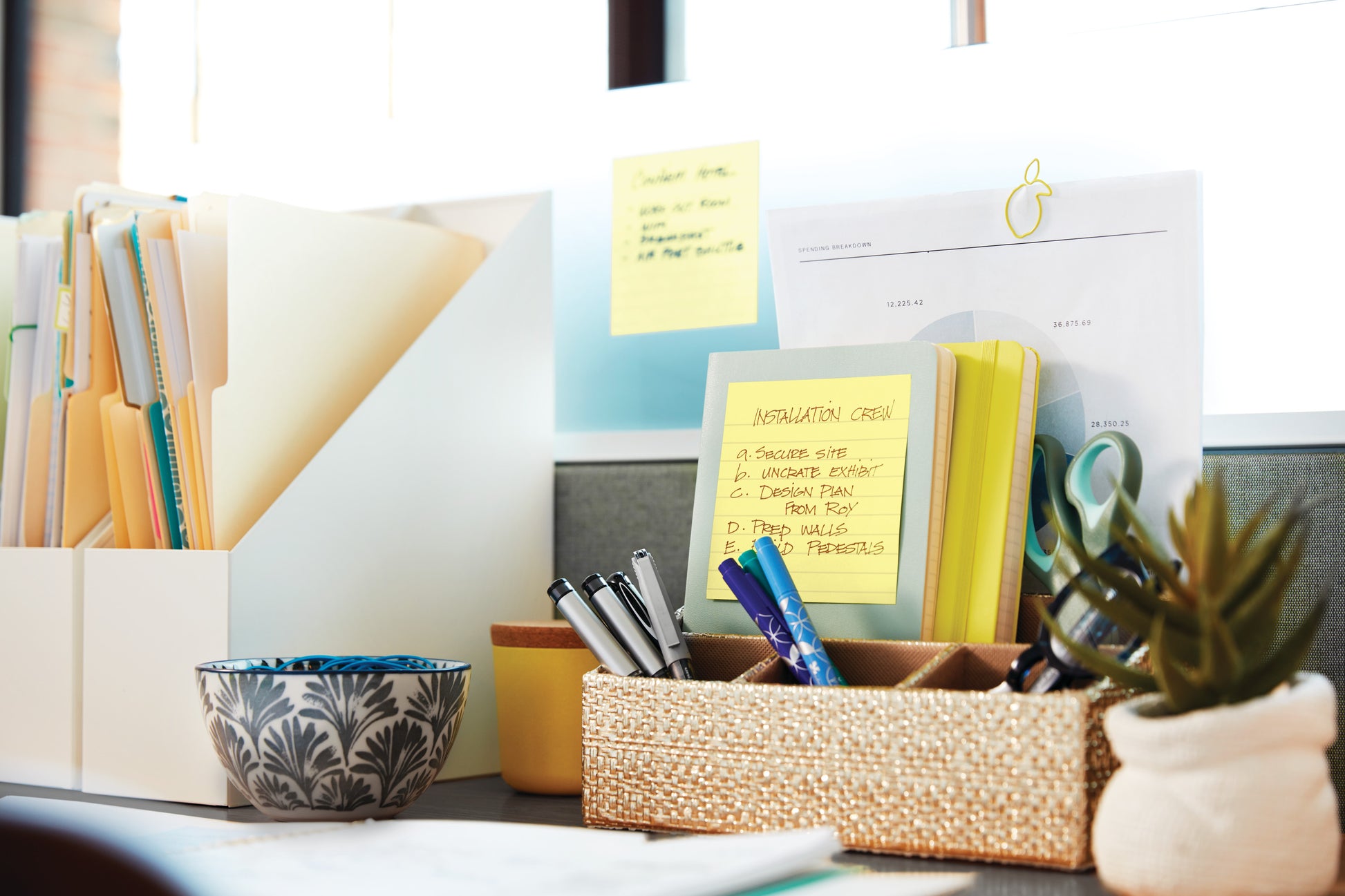 Ein aufgeräumter Schreibtisch mit 3M Post-it® Super Sticky Notes (101x101 mm, 90 Blatt, 100% PEFC), Aktenordnern, einer gemusterten Schale, einem geflochtenen Korb für Stifte und Scheren und Papieren am Fenster.