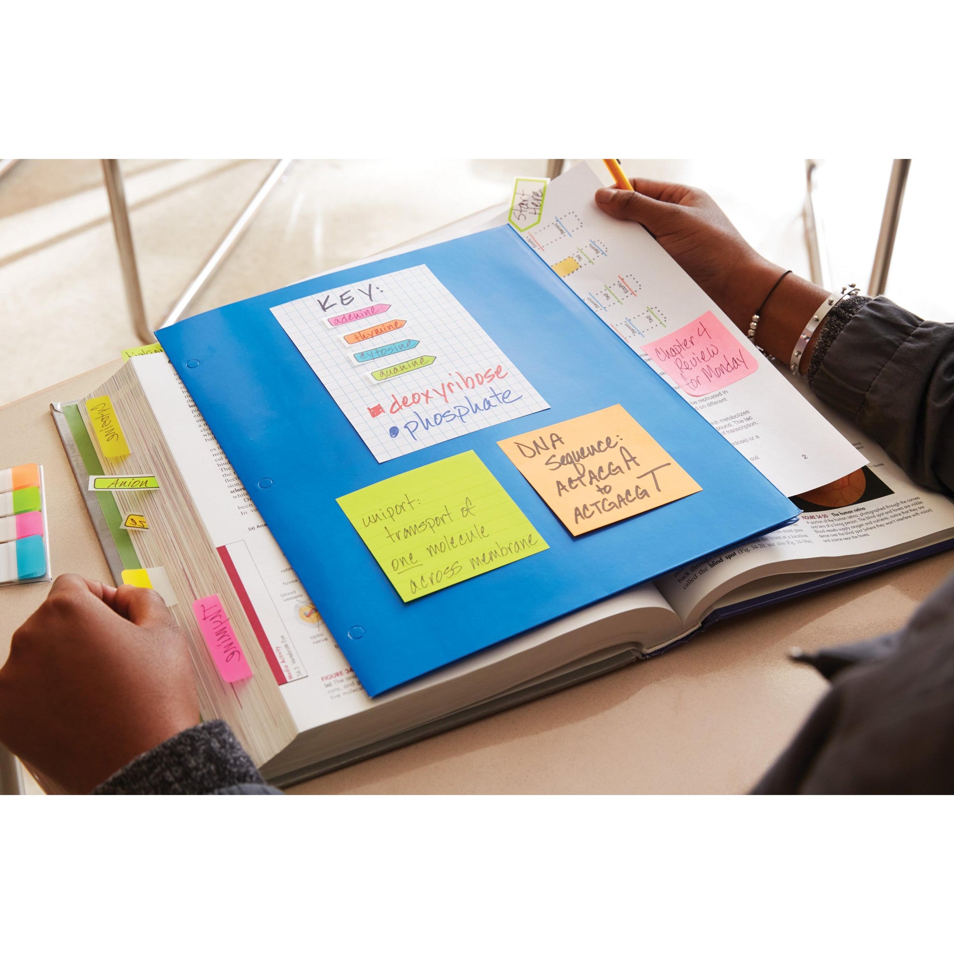 Ein Student studiert ein Biologie-Lehrbuch, das mit 3M Deutschland GmbH Post-it® Notes, Energetic Collection, beklebt ist. Er notiert Genetik und Zellmembranen auf den Haftnotizen (76 mm x 127 mm), während die Lernmaterialien auf dem Schreibtisch verstreut liegen.