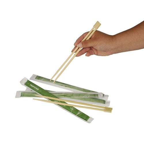 Eine Hand hält PAPSTAR 50 Paar Essstäbchen „pure“ 23 cm, jedes Paar hygienisch verpackt in grünen Hüllen der PAPSTAR GmbH, darüber mehrere weitere Paare auf weißem Untergrund.