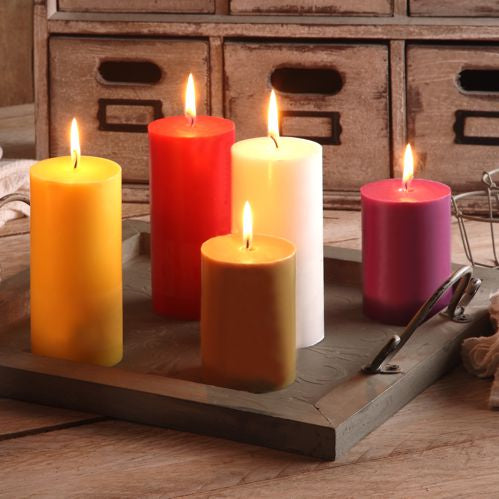 Fünf brennende PAPSTAR Stumpenkerzen Ø 70 mm, 100 mm durchgefärbte Kerzen der PAPSTAR GmbH aus 100 % Stearin in Gelb, Rot, Weiß, Braun und Rosa stehen auf einem Holztablett mit Griffen auf einem rustikalen Tisch vor kleinen Holzschubladen.