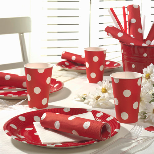 Ein Tisch ist mit roten Papptellern, Bechern und 20 PAPSTAR Servietten (40x40 cm, 3-lagig) der PAPSTAR GmbH gedeckt, alle mit weißen Punkten. Weiße Gänseblümchen schmücken den Tisch, während das Licht durch weiße Jalousien im Hintergrund fällt.