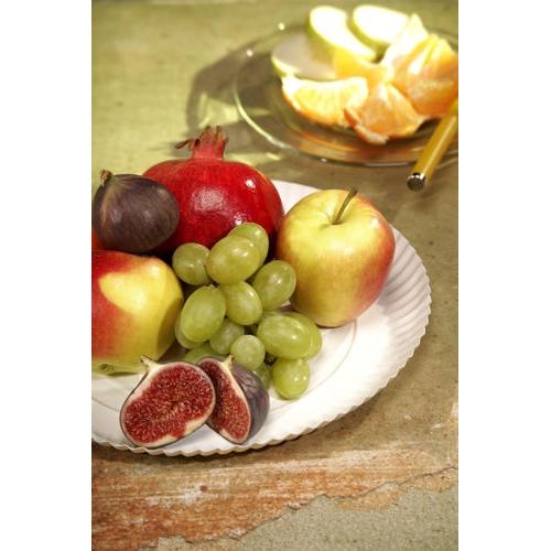 A PAPSTAR 100 plates, cardboard "pure" round from PAPSTAR GmbH holds apples, green grapes, figs, and a pomegranate. In the background rests a glass plate with orange slices and a knife with a yellow handle on a greenish surface.