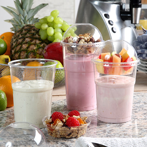 Drei Smoothies – einer pur, einer pink, einer mit Früchten und Granola – werden in PAPSTAR 60 Kaltgetränkebechern PLA „pure“ (0,5 l) serviert. Präsentiert werden sie auf einer Theke, in deren Nähe sich Früchte, ein Mixer sowie Beeren und Granola befinden. Marke: PAPSTAR GmbH.