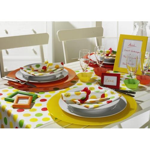 Ein Tisch für zwei Personen mit gelben und orangefarbenen Platzdeckchen, weißen Tellern, PAPSTAR 20 Servietten 33x33 cm von PAPSTAR GmbH, Gläsern, gerahmten Tischkarten, einer rot-gelb-grün gepunkteten Tischdecke und Dekoration sowie einer gerahmten Speisekarte im Hintergrund.