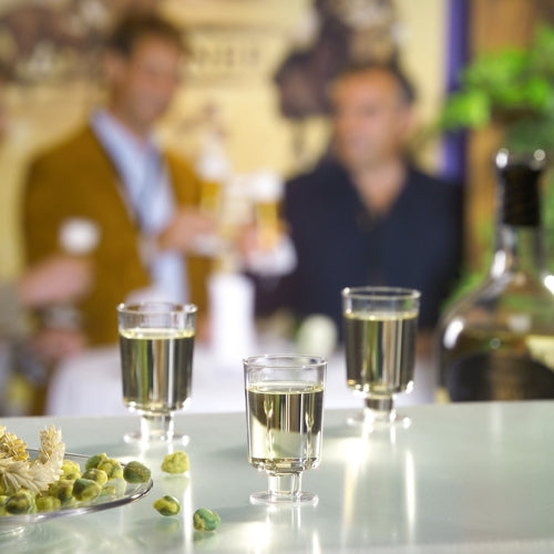 Die Starpak 20 Stiel-Gläser für Weißwein aus glasklarem Polystyrol stehen auf einem Tisch mit Oliven und Flasche, während sich drei Personen im Hintergrund bei Getränken unterhalten.