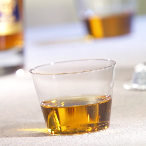 A Starpak 15 drinking cup, PS 0.1 l crystal clear, contains a small amount of amber liquid on a light surface, while another cup and a bottle are blurred in the background.