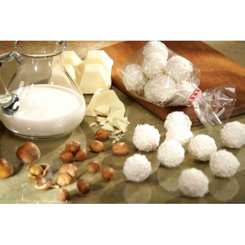 Auf einer Arbeitsplatte sind ein Glaskrug mit Milch, weißen Schokoladenstückchen, Haselnüssen und mit Kokosnuss überzogenen Bonbonkugeln angeordnet, einige Leckereien werden in einem Starpak 300 Bodenbeutel (transparentes PP) von Starpak auf einem hölzernen Schneidebrett präsentiert.