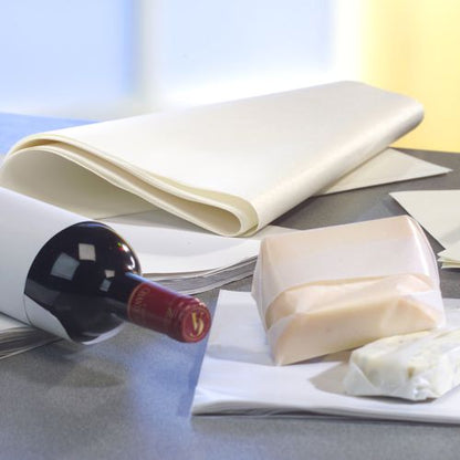 A bottle of red wine and cheese blocks are partially wrapped in PAPSTAR 10 kg wrapping papers, white cellulose from PAPSTAR GmbH, in the background a stack of wax paper sheets can be seen on the tabletop.