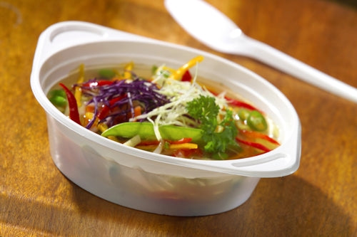 Eine Starpak 100 Suppenschalen, PP rund, Ø 15,6 cm, enthält eine kräftige Gemüsesuppe mit roter Paprika, Kohl, Erbsen und Petersiliengarnitur auf einem Holztisch neben einem weißen Plastiklöffel.