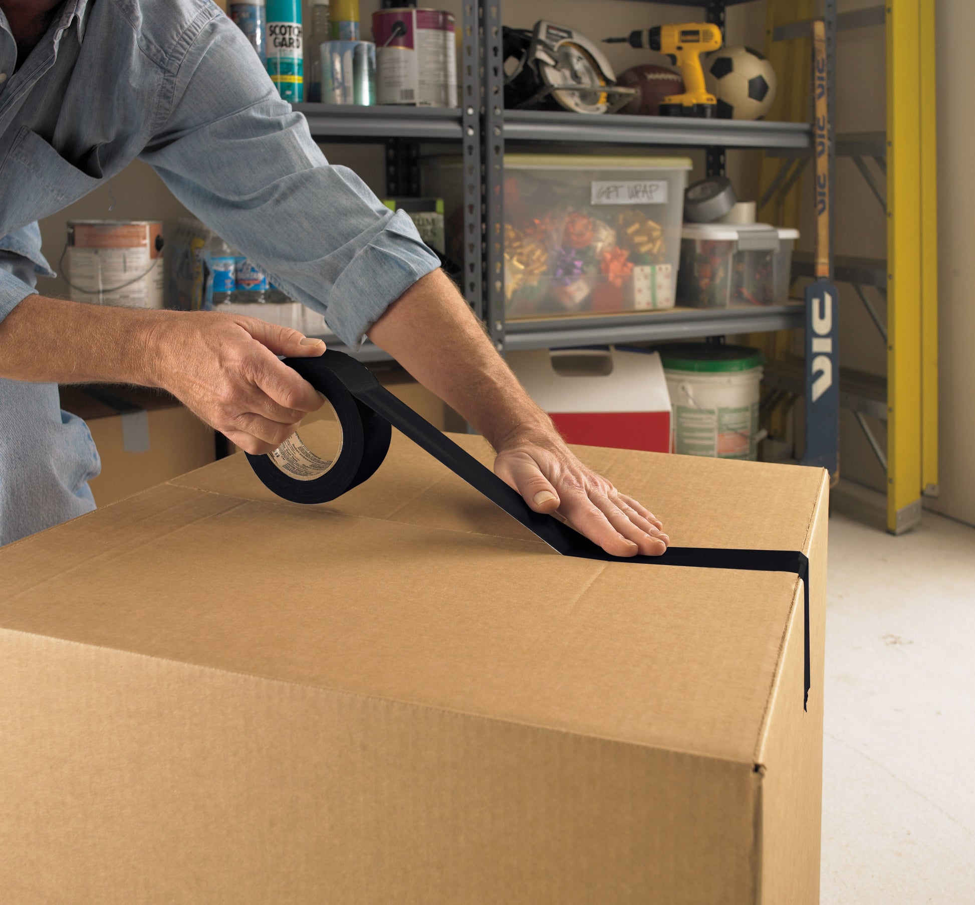 Eine Person verschließt einen großen Karton mit Scotch® Universal Klebeband 2904 (10 m x 48 mm) von 3M Deutschland GmbH in einer Garage. Im Hintergrund sind Regale mit Werkzeug, Farbdosen und Haushaltsgegenständen zu sehen.