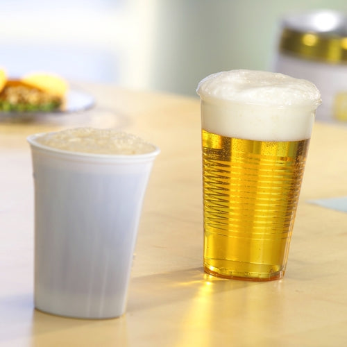 Zwei Bierbecher stehen auf einem hellen Holztisch - einer in einem klaren Starpak 75 Trinkbecher, PS klar mit Schaumrand von Starpak, der goldenes Bier zeigt, der andere in einem undurchsichtigen weißen Becher. Im Hintergrund sind unscharfe Lebensmittel zu sehen.