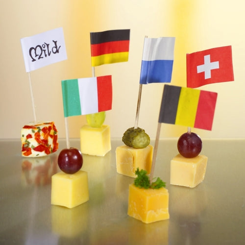 Sechs Käsewürfel sind mit Trauben oder Kräutern belegt und für eine stilvolle und umweltfreundliche Präsentation mit den 8 cm großen weißen Deko-Picker-Fähnchen der PAPSTAR GmbH geschmückt, wobei auf jedem Fähnchen „Mild“, Deutschland, Italien, Finnland, die Schweiz oder Belgien steht.