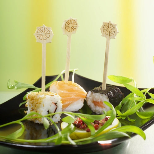 Drei Sushistücke, jeweils mit einem Fingerfoodpicker „Steuerrad“ (9 cm) der PAPSTAR GmbH aus der 200-Stück-Packung, werden auf einem schwarzen Teller mit grüner Garnitur und Gurke mit roten Beeren serviert, vor einem gelb-grünen Farbverlaufshintergrund.