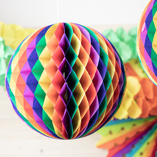 Eine Nahaufnahme des PAPSTAR Wabenballs „schwer entflammbar“ der PAPSTAR GmbH, einer kugelförmigen Dekoration in Regenbogenfarben, mit anderem farbenfrohen Dekor, das im Hintergrund sanft verschwommen ist.