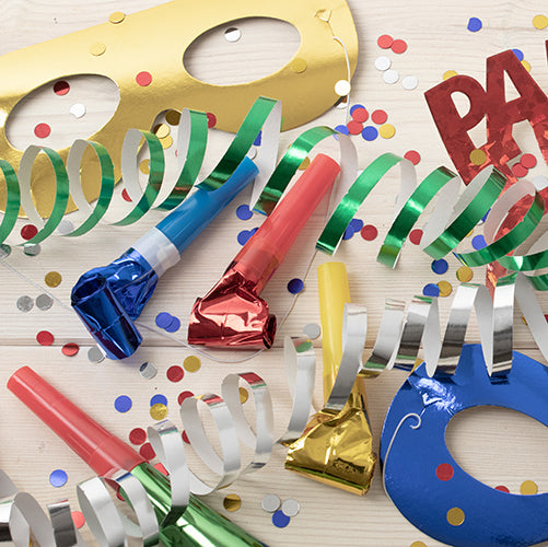 Die 6 Gesichtsmasken farbig sortiert „Metallic“ (6 Stück) der PAPSTAR GmbH plus Krachmacher, Metallic-Farben-Luftschlangen und Konfetti sind auf einer hellen Holzoberfläche angeordnet.