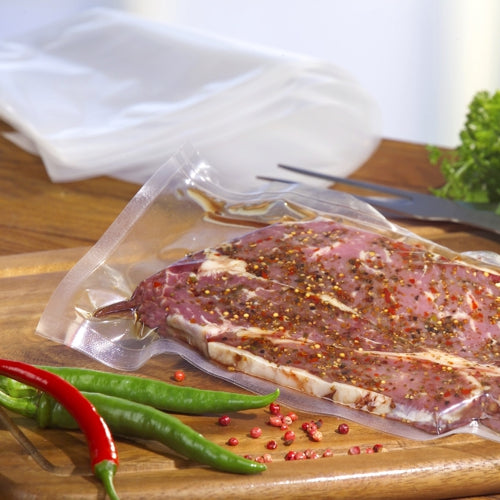 Ein Starpak 100 Siegelrandbeutel, PA / PE transparent, gefüllt mit mariniertem Fleisch liegt auf einem hölzernen Schneidebrett inmitten von Chilischoten und rosa Pfefferkörnern. Blattgemüse und Utensilien vervollständigen die Szene - ideal für die Verwendung mit einem Kammervakuumierer.