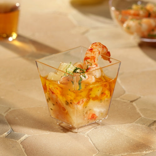 Eine Starpak 50 Fingerfood-Schale (PS glasklar) von Starpak, gefüllt mit Garnelen und Meeresfrüchtecocktail und garniert mit Kräutern, steht auf einem beigefarbenen, strukturierten Untergrund. Weitere Tassen sind im Hintergrund verschwommen – perfekt für stilvolle Events.