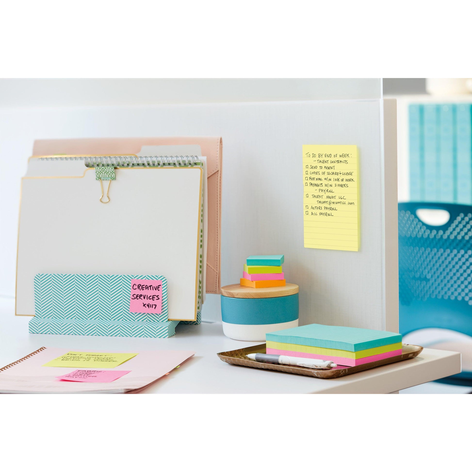 Ein aufgeräumter Bürotisch mit Aktenordnern, einem Notizblock, bunten Haftnotizen, einem Stift, einem Tablett und einem runden Behälter. Auf der Trennwand liegt ein gelbes Post-it® Notes im Großformat (101 mm x 152 mm) der 3M Deutschland GmbH, im Hintergrund stehen blaue Stühle und Schränke.