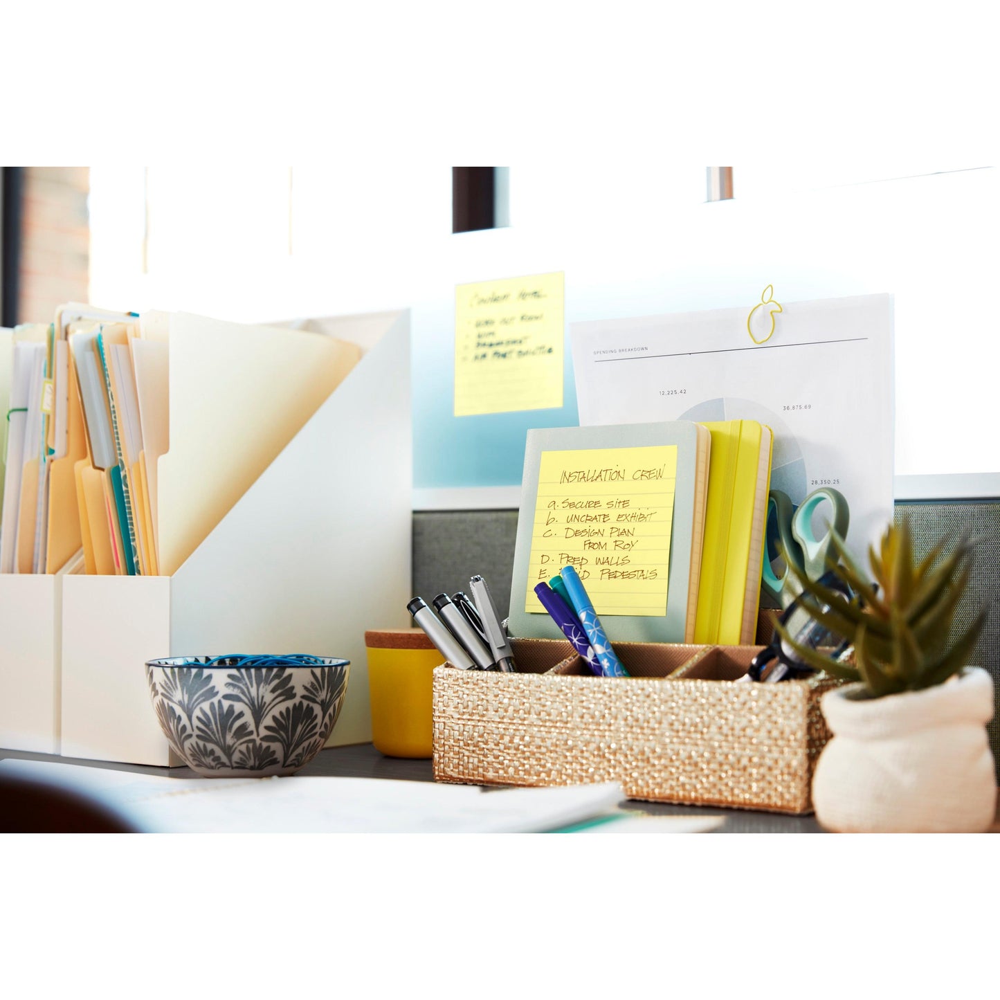 A tidy desk with folders, documents, pens in a woven container, a decorative bowl, a small plant, and 3M Deutschland GmbH Post-it® Notes in large format (Yellow, 101x152 mm), one with the inscription "Installation Crew," at a sunny window.