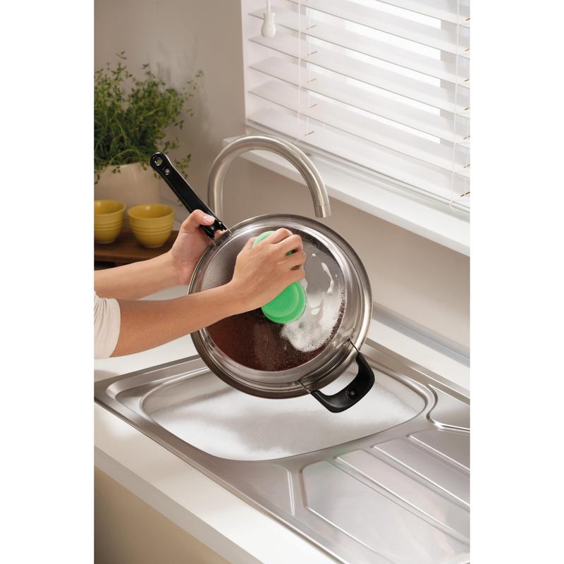 Someone is cleaning a metal pot lid over a soapy sink with the Scotch-Brite® Classic Cleaning Sponge from 3M Deutschland GmbH, near a window with white blinds and a counter with a plant and stacked yellow bowls.
