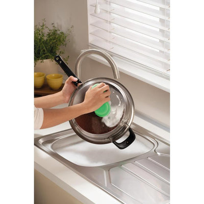 Someone is cleaning a metal pot lid over a soapy sink with the Scotch-Brite® Classic Cleaning Sponge from 3M Deutschland GmbH, near a window with white blinds and a counter with a plant and stacked yellow bowls.
