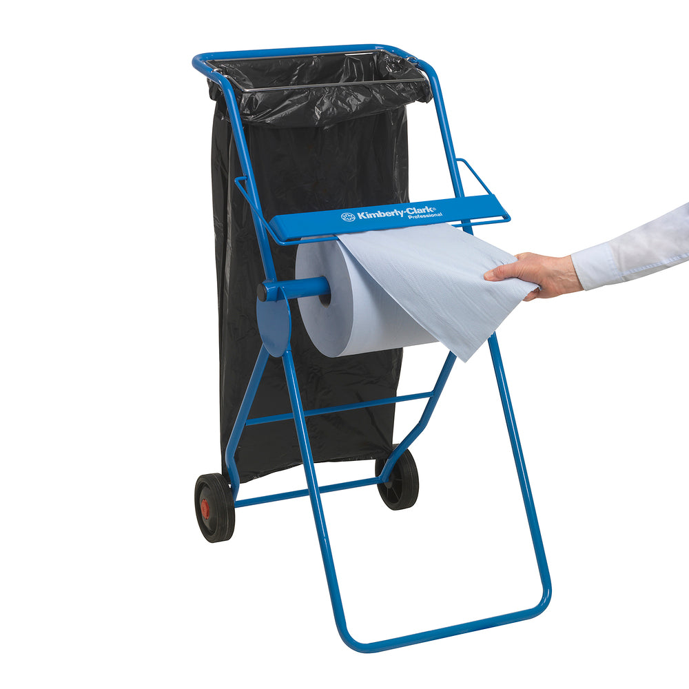 Ein blauer Metallständer mit zwei Rädern hält einen schwarzen Müllsack und eine WypAll® L10 EXTRA Wischtücher Großrolle (1 Rolle x 1500 Blatt); eine Person zieht ein Blatt von der Rolle. Auf der horizontalen Fläche des Ständers ist die Aufschrift "Kimberly-Clark GmbH" angebracht.