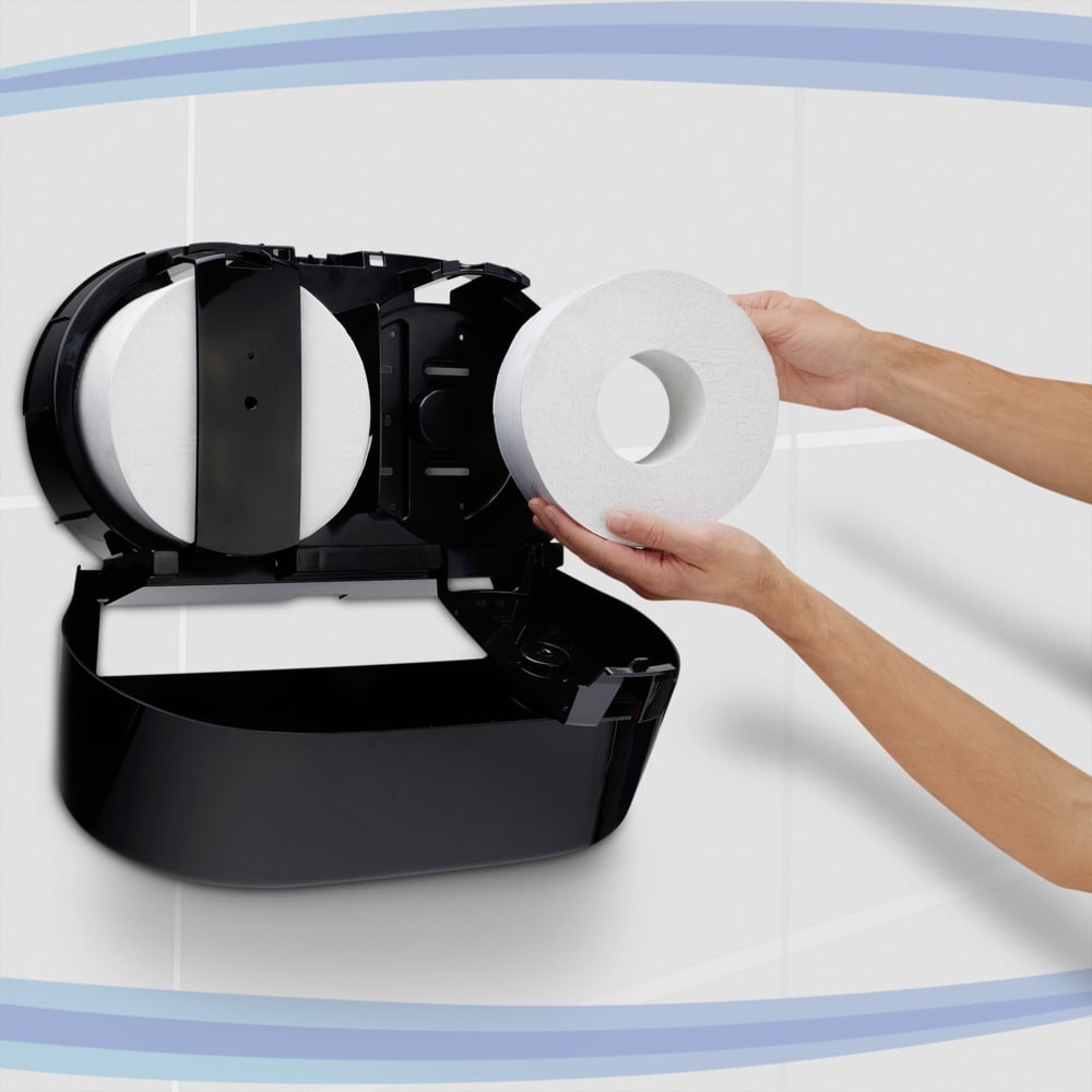 Eine Person lädt eine Jumbo-Toilettenpapierrolle in einen schwarzen Kimberly-Clark Professional™ Toilet Tissue Spender - Zentralentnahme von Kimberly-Clark GmbH, der an einer gekachelten Wand mit blauen geschwungenen Linien im Hintergrund angebracht ist.