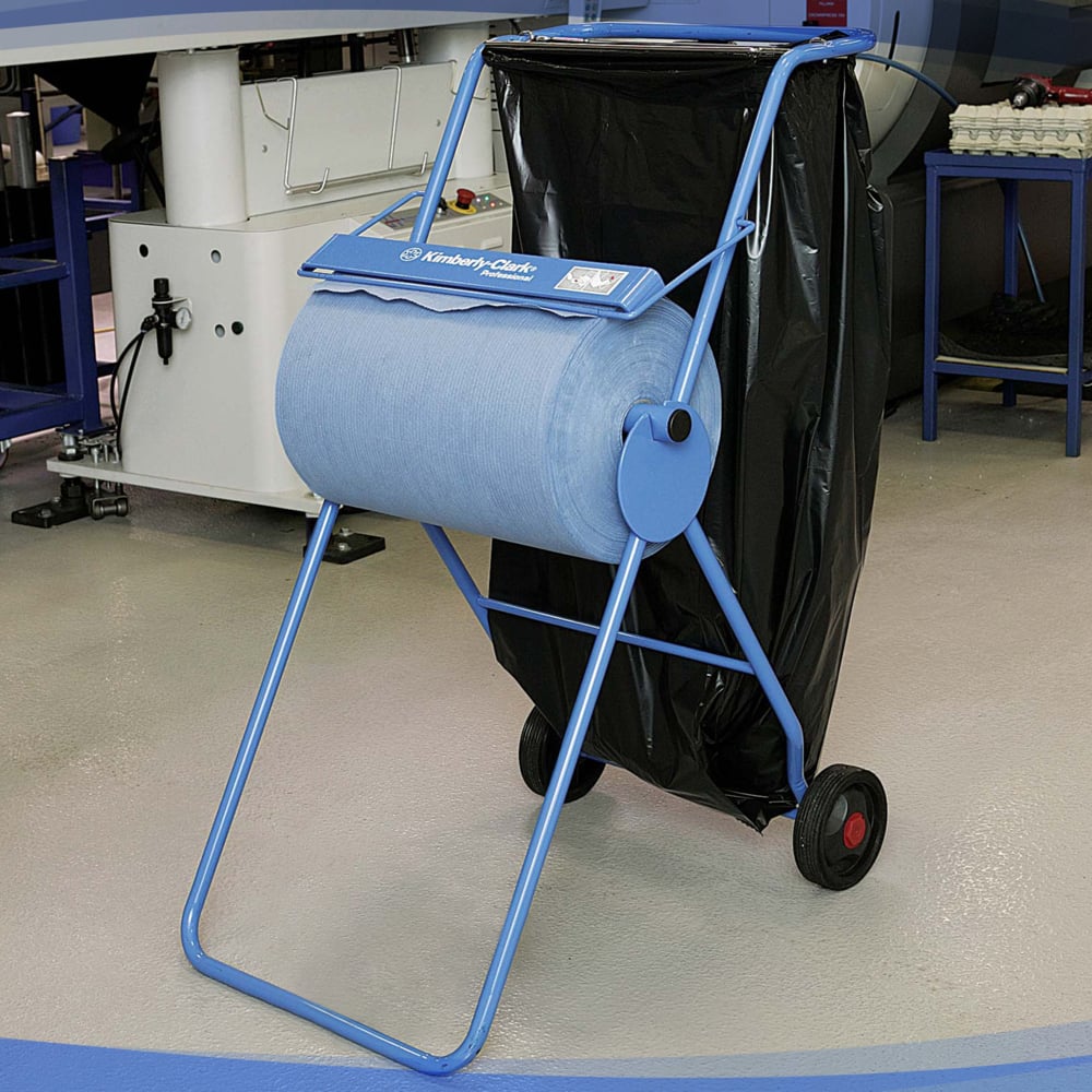 Ein Kimberly-Clark Professional™ Mobiler Ständer für Wischtuchrollen (Großrolle/Blau) mit einer Rolle blauer Industriepapierhandtücher und einem großen schwarzen Müllsack steht auf Rädern auf einem grauen Hallenboden an einem Arbeitsplatz.