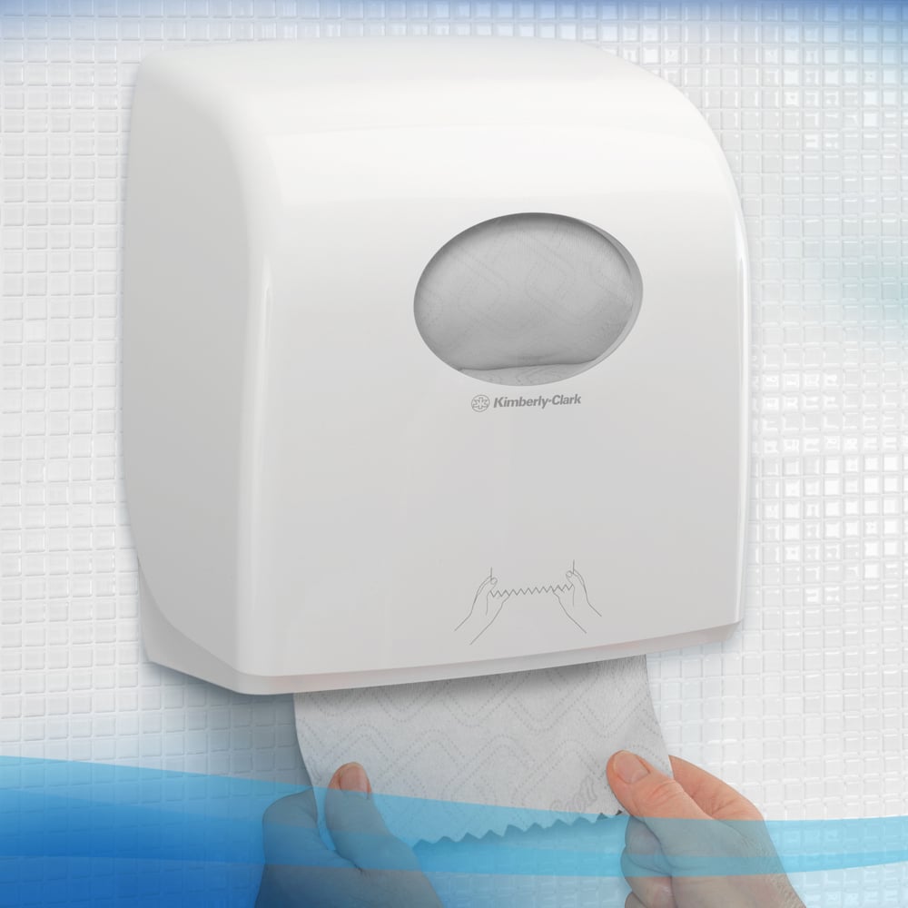 A person pulls a towel from a white Kimberly-Clark GmbH Scott® Essential™ towel dispenser with a small oval window through which the paper roll inside can be seen, with textured tiles in the background. 6 white rolls per box.