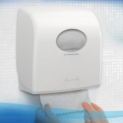 A person pulls a towel from a white Kimberly-Clark GmbH Scott® Essential™ towel dispenser with a small oval window through which the paper roll inside can be seen, with textured tiles in the background. 6 white rolls per box.