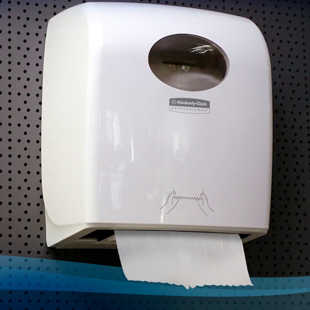 Ein weißer Wandspender mit transparentem Sichtfenster hält eine Scott® Handtücher - Rolle (304,00 m x 19,80 cm) der Kimberly-Clark GmbH mit einem herunterhängenden Blatt auf einer gelochten grauen Wand.