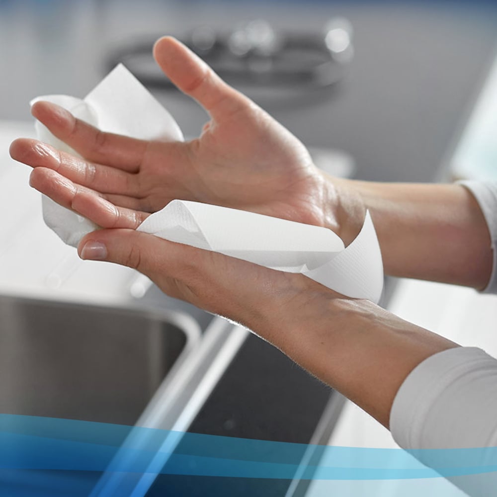 Eine Person wischt sich die Hände mit einer weißen Scott® Handtücher - Rolle (304,00 m x 19,80 cm) von Kimberly-Clark GmbH über einem Spülbecken ab, wobei die Arbeitsplatte und das Spülbecken im Hintergrund zu sehen sind.