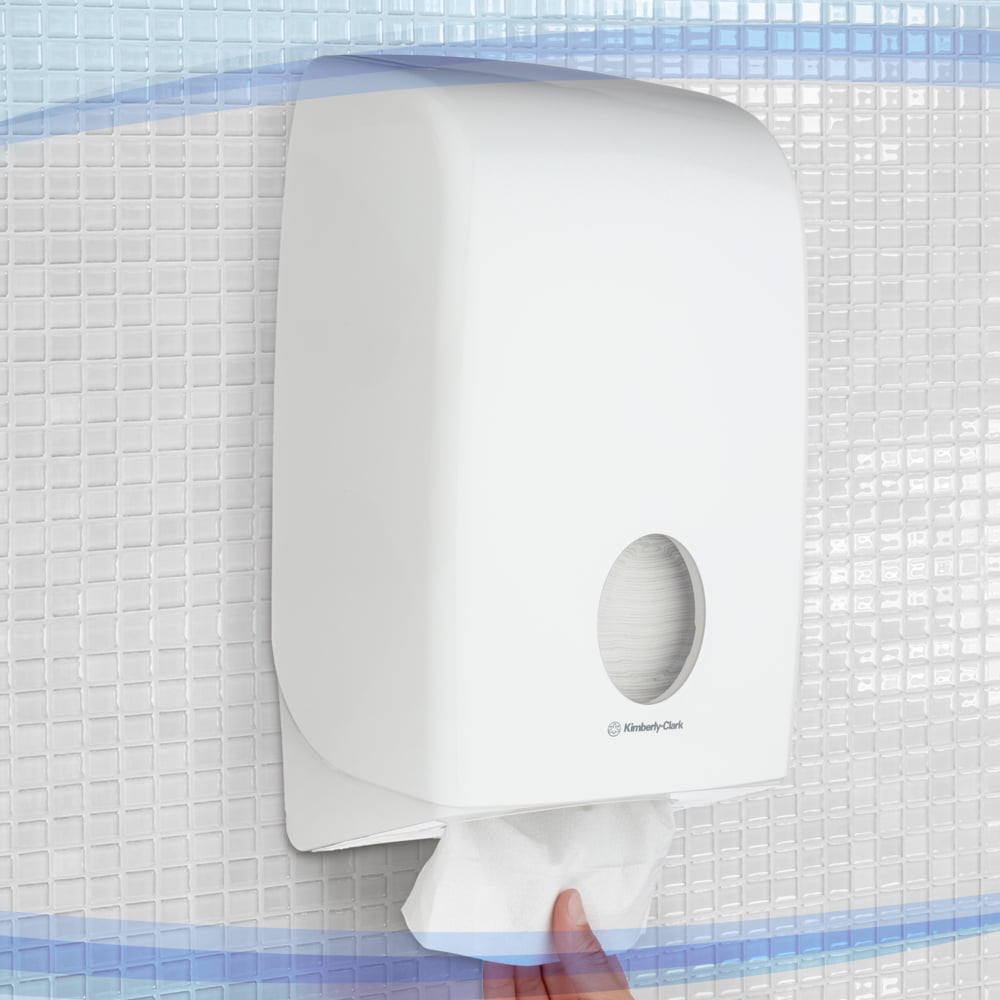 A hand pulls a paper towel from the white Aquarius™ dispenser for folded towels from Kimberly-Clark GmbH, mounted on a tile on the wall. The dispenser has a window for hygienic, easy towel dispensing.