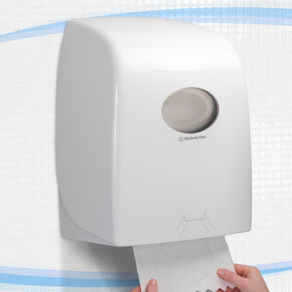 The Aquarius™ roll towel dispenser - White by Kimberly-Clark GmbH is wall-mounted on tiles, with a hand pulling a towel from below. It is ideal for public areas and features a round window at the top and clear usage instructions below.