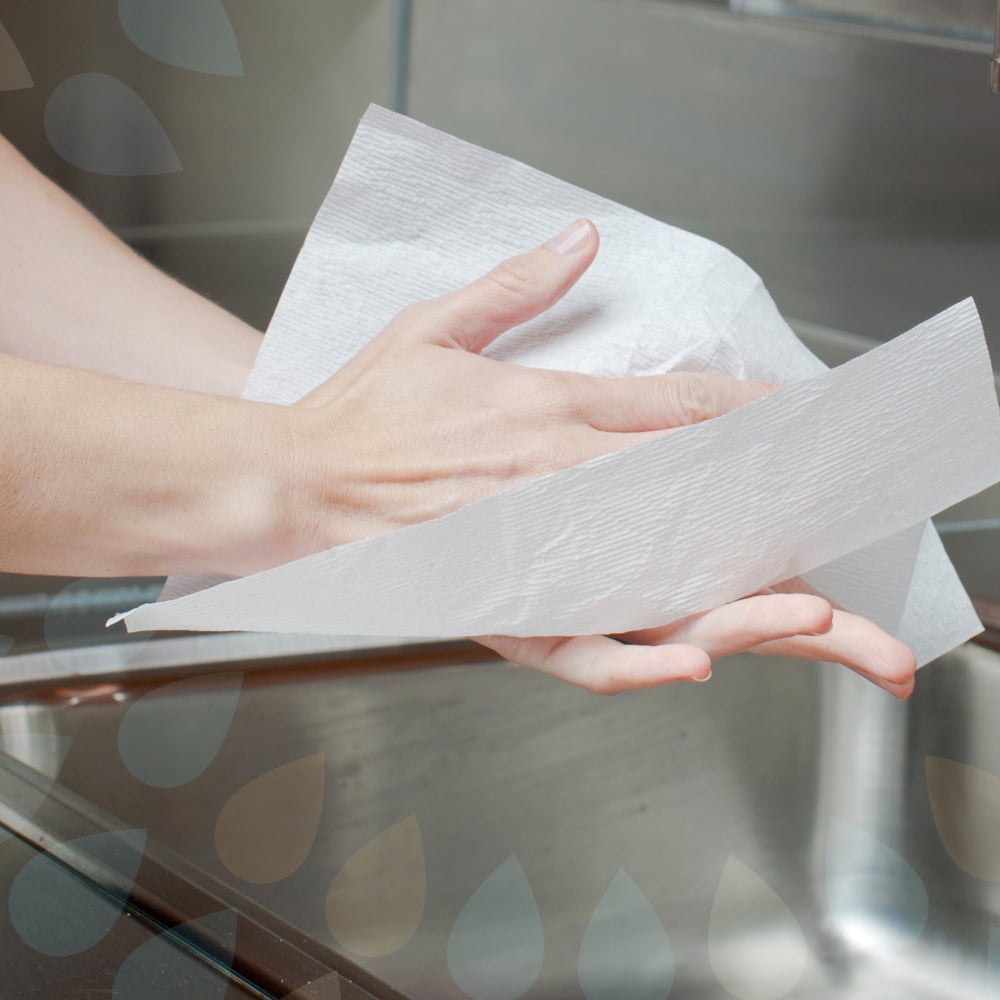 Eine Person trocknet sich die Hände mit einem weißen Kleenex® Ultra™ Handtücher Interfold Papierhandtuch der Kimberly-Clark GmbH über einem Waschbecken aus Edelstahl, um eine hygienische Erfahrung zu machen.