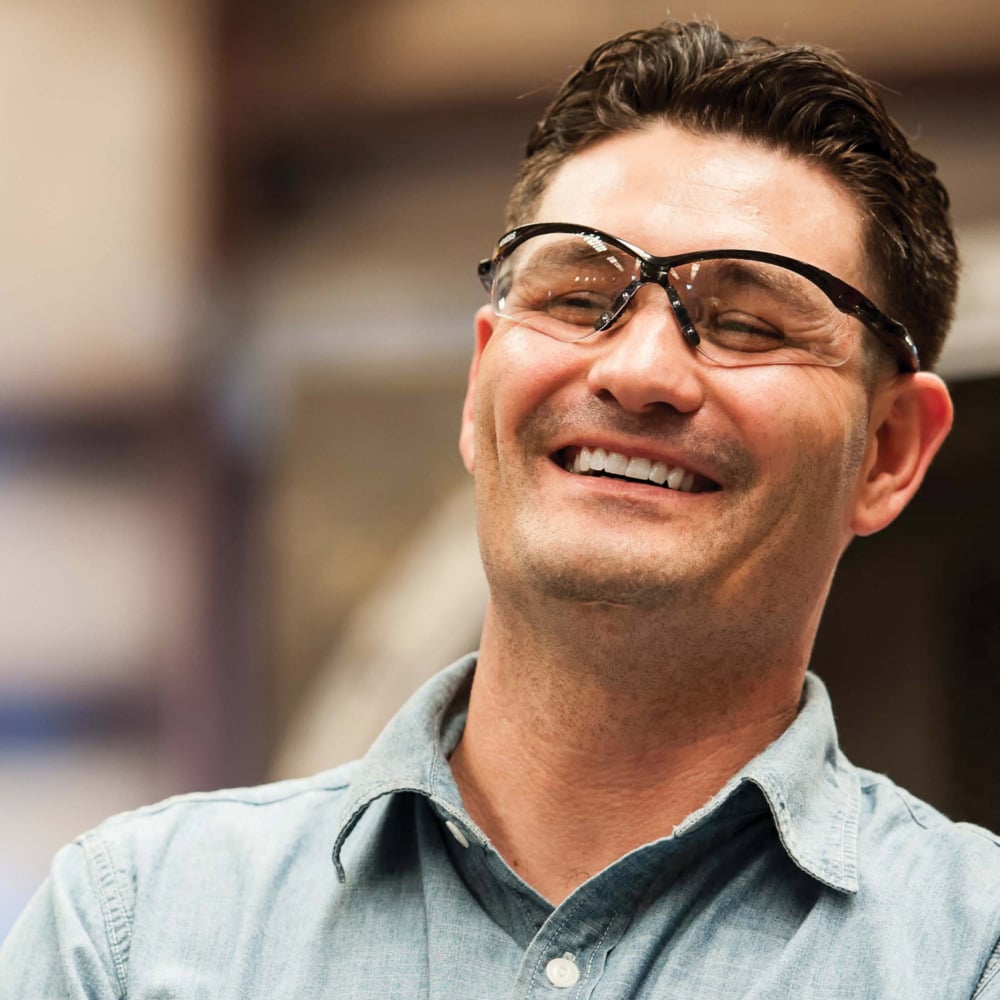 Ein Mann in einem hellblauen Hemd lächelt, während er die KleenGuard® V30 Nemesis™ Schutzbrille mit beschlagfreier Beschichtung von Kimberly-Clark GmbH trägt. Der unscharfe Hintergrund hebt ihn und seine Schutzbrille hervor.