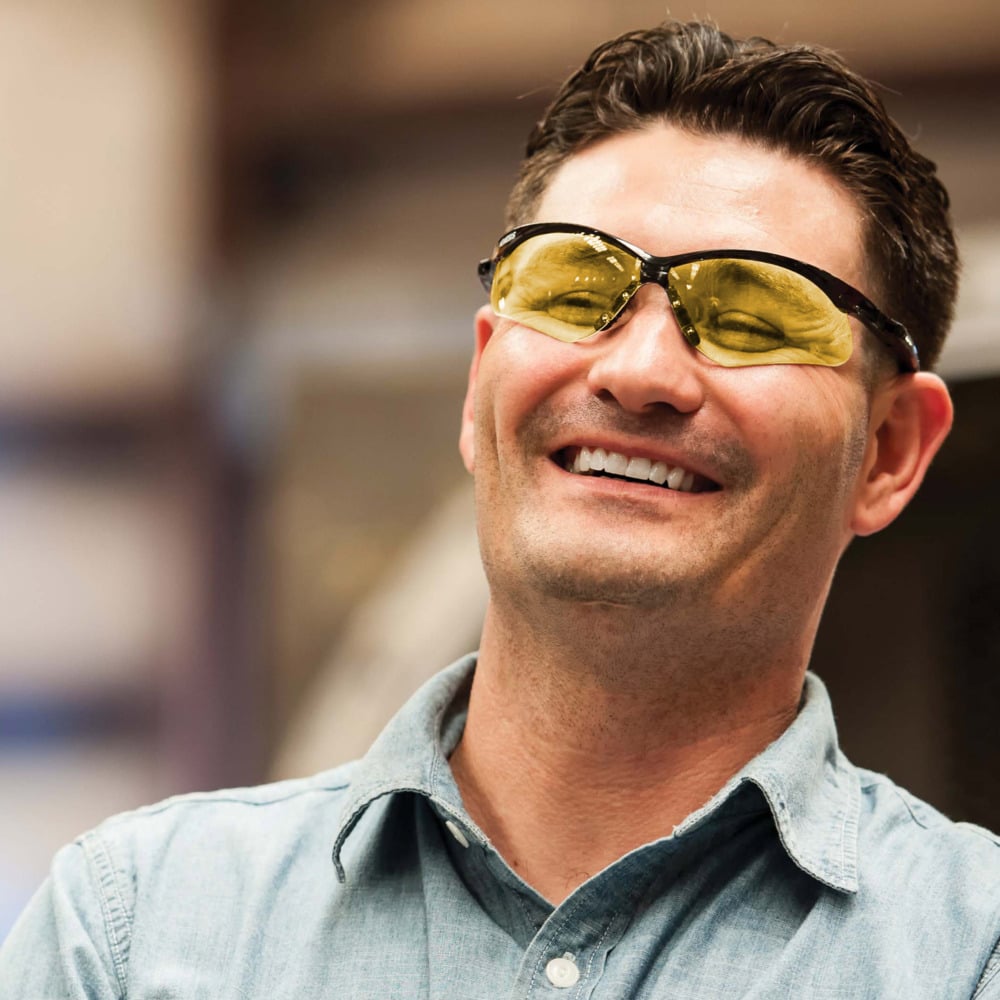 Ein Mann im hellblauen Kragenhemd und KleenGuard® V30 Nemesis™ Schutzbrille - Caramel von Kimberly-Clark GmbH lächelt. Die gelb getönte Brille sorgt für optimalen Augenschutz. Der Hintergrund ist unscharf.