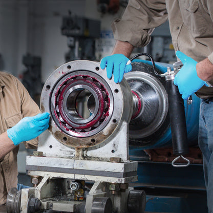 Zwei Personen in hellbraunen Hemden tragen Kimberly-Clark GmbH KleenGuard® G10 Comfort Plus Blaue Nitril-Handschuhe (24 cm, beidhändig tragbar), während sie mit einer Fettpresse Schmiermittel auf ein Lager einer Industrieanlage in einer Werkstatt auftragen.