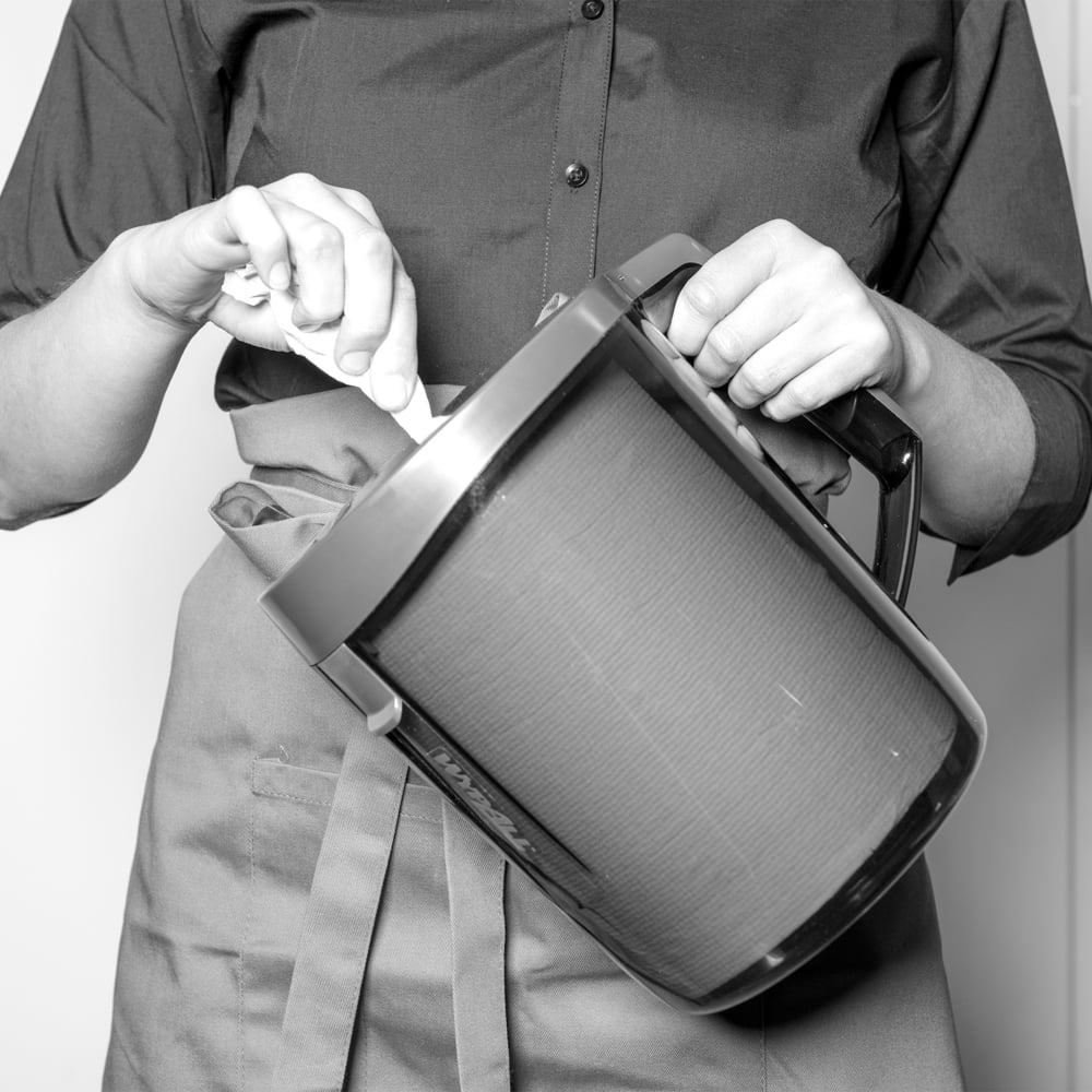 Eine Person in Schürze und geknöpftem Hemd rührt einen großen Krug um, während ein WypAll® Reach™ Centrefeed Dispenser (Schwarz) der Kimberly-Clark GmbH in der Nähe steht und eine professionelle Note verleiht. Das Schwarz-Weiß-Bild hebt die Szene hervor.