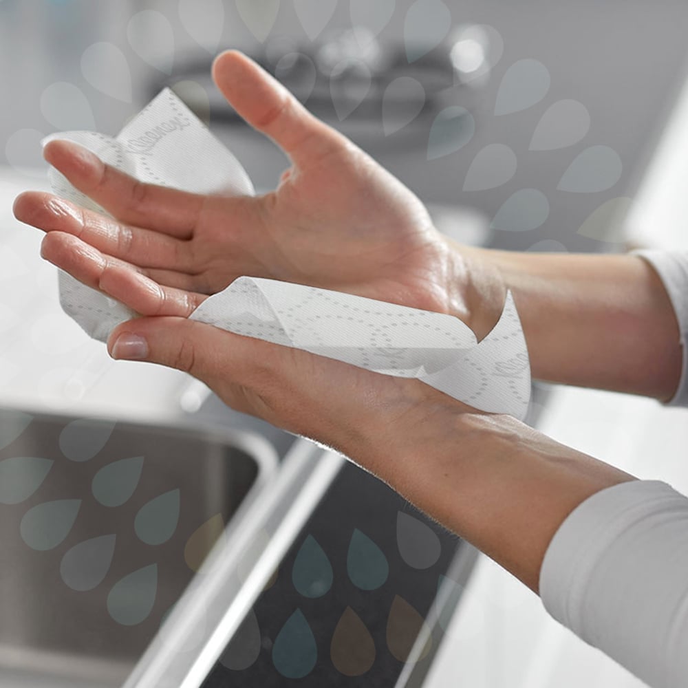 A person dries their hands near a sink with Kleenex® roll towels E-roll Large (white, 1-ply, 6x250m) from Kimberly-Clark GmbH. Translucent water droplets in the background emphasize the hygienic hand drying.